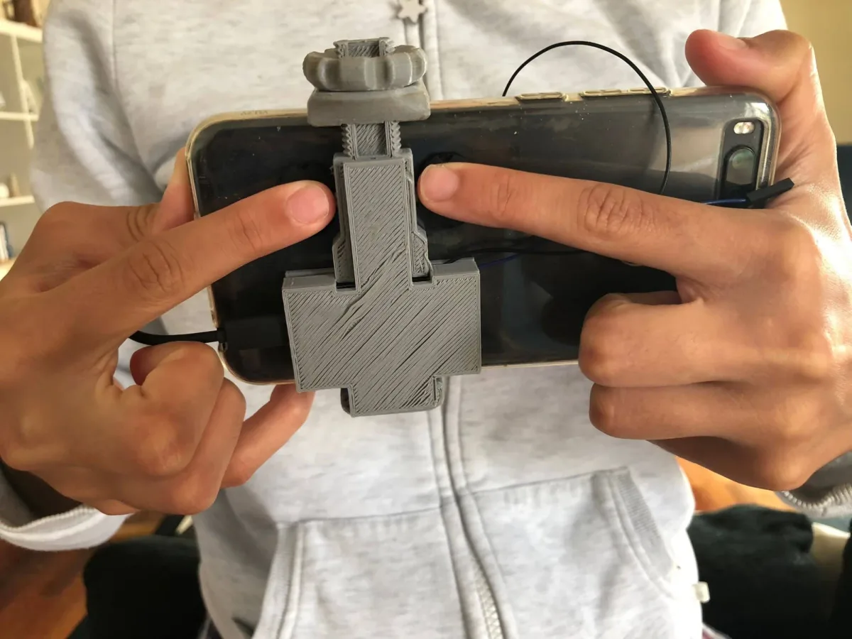 A phone held in two hands, both index fingers resting on a printed module clamped to the back of the case.