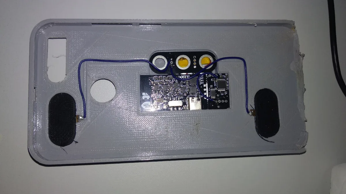 The inside of a printed phone case: dark fabric electrode pads at each end, blue wires running to a circuit board with a USB-C connector and a smaller board beside it.