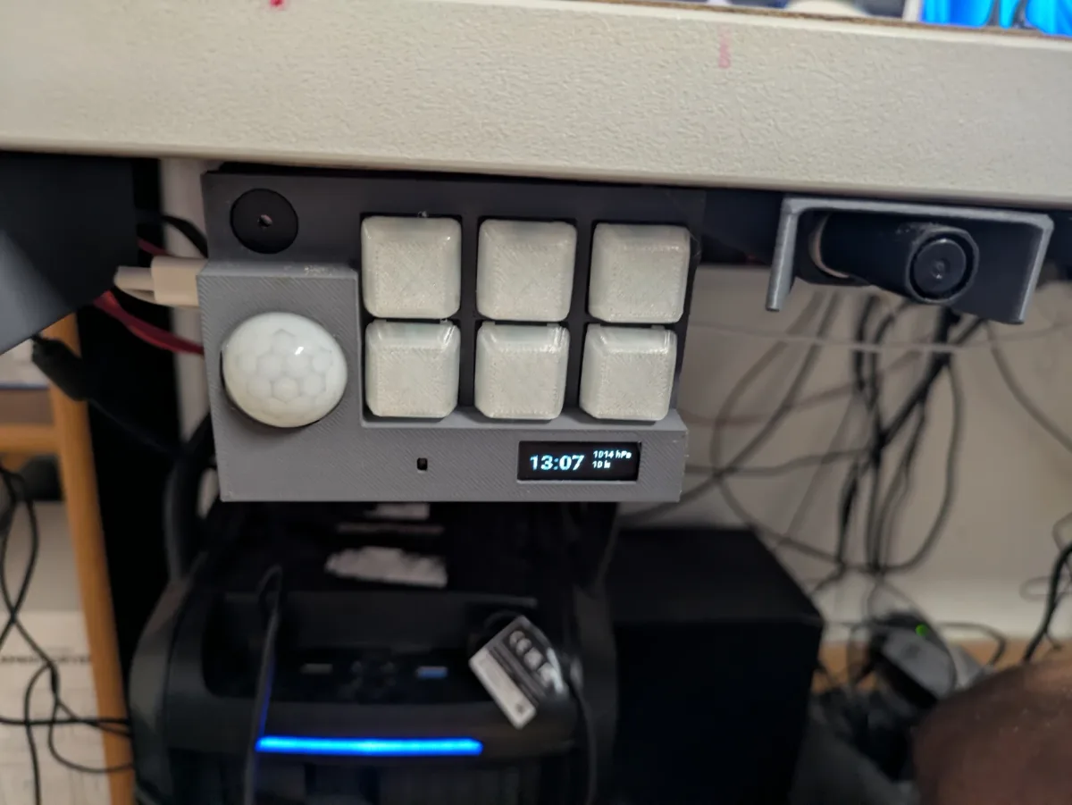 A grey printed enclosure mounted under the edge of a desk, with six white keycaps, a PIR sensor dome, a buzzer, and a small OLED showing the time, air pressure and light level.