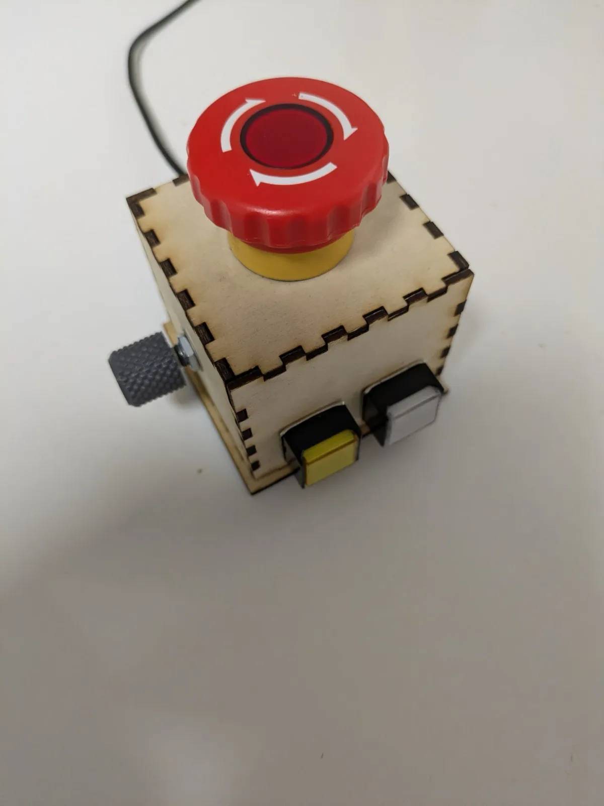 A small laser-cut plywood box with finger-jointed corners, a large red latching mushroom button on top, two switches on the front face and a knurled thumbscrew on the side.