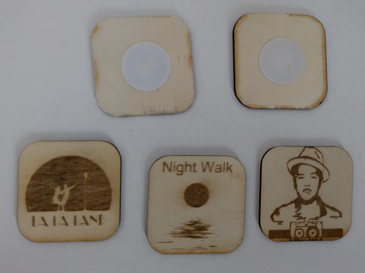 Five laser-cut plywood squares. Two lie face down showing white circular NFC stickers; three are engraved with album artwork.