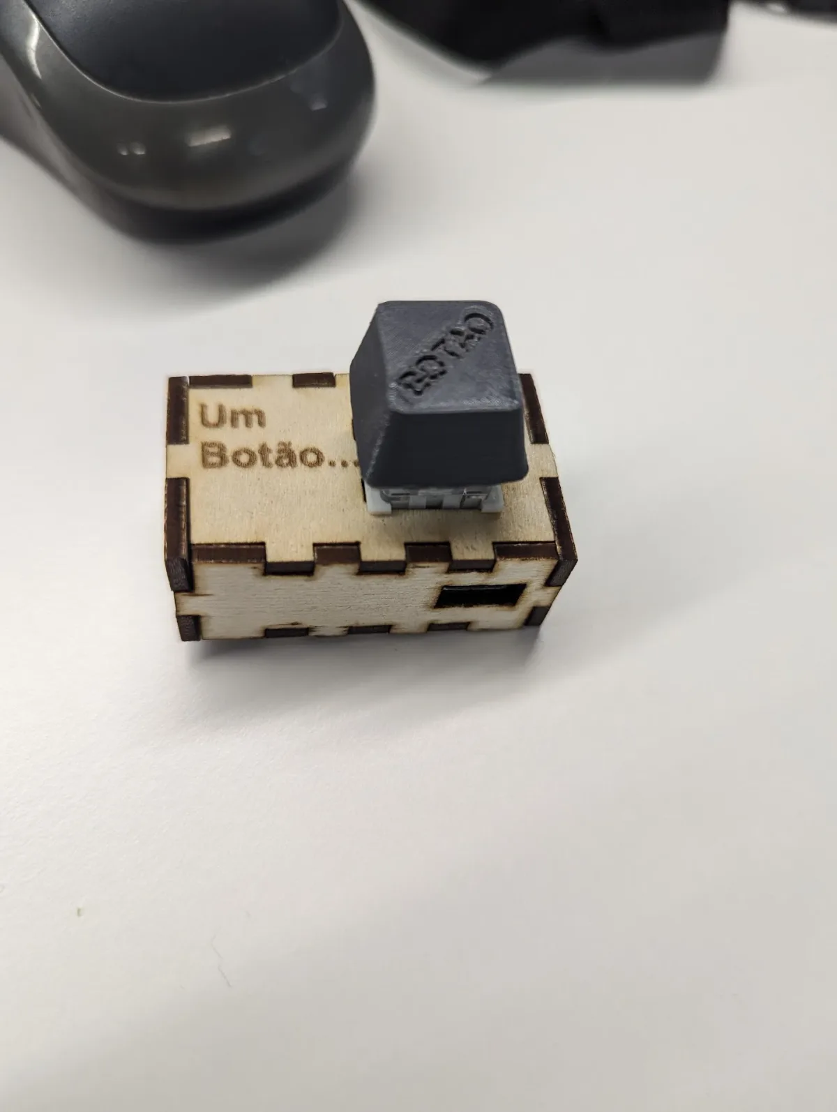 A small finger-jointed plywood box engraved with the words Um Botão, topped by an oversized 3D-printed keycap embossed BOTÃO on a mechanical switch.
